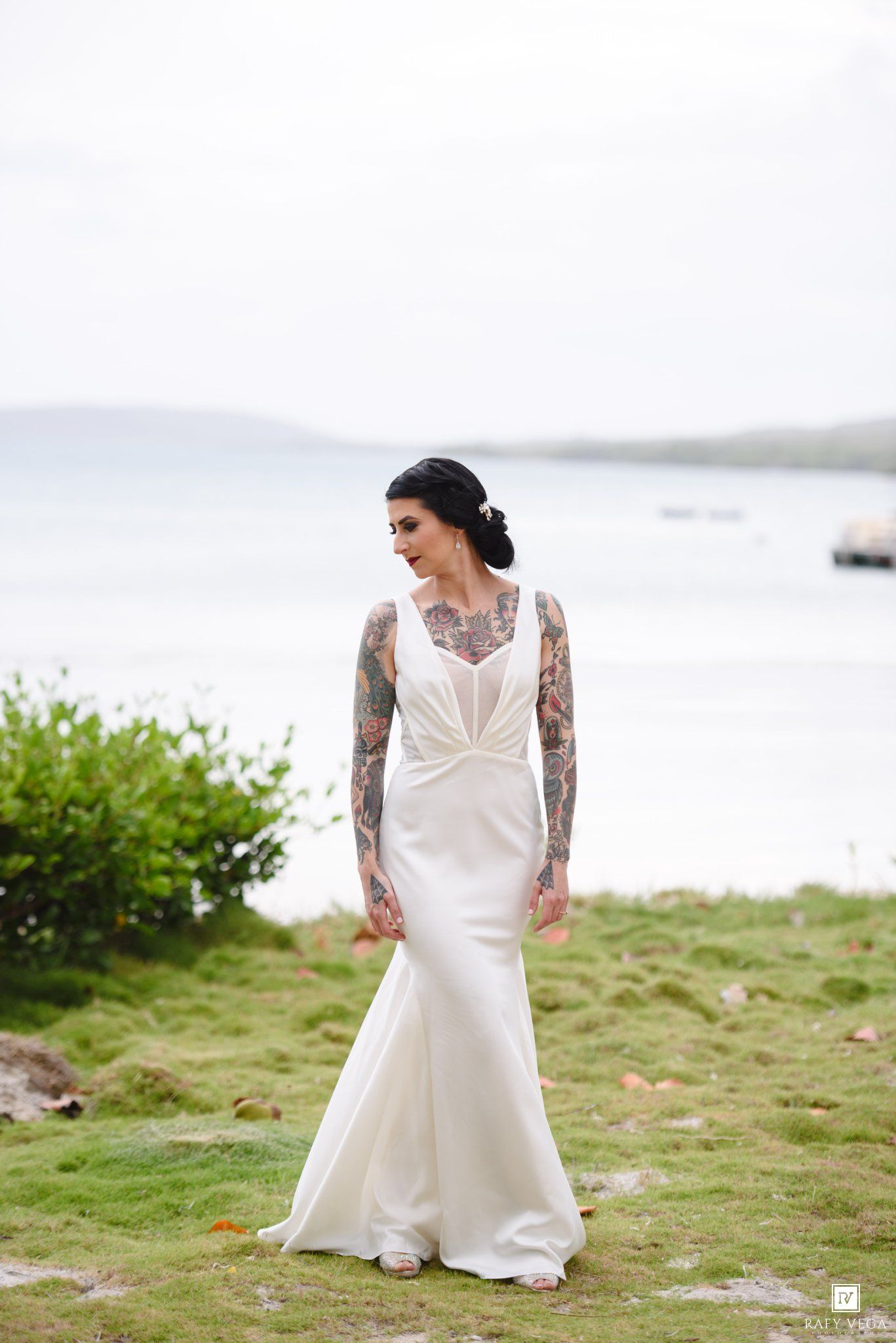 Tattooed bride in elegant white mermaid wedding gown standing on coastal grass with ocean views in background.