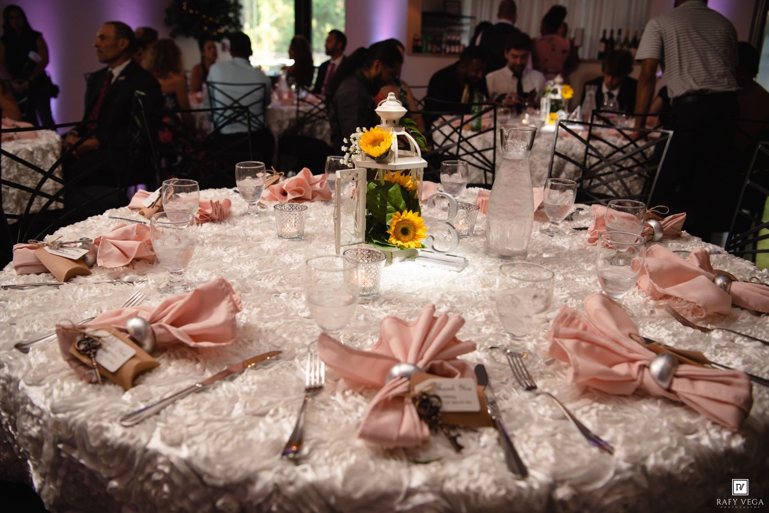 Elegant wedding reception table with pink satin napkins, white lace tablecloth, and sunflower centerpiece in a warmly lit venue.