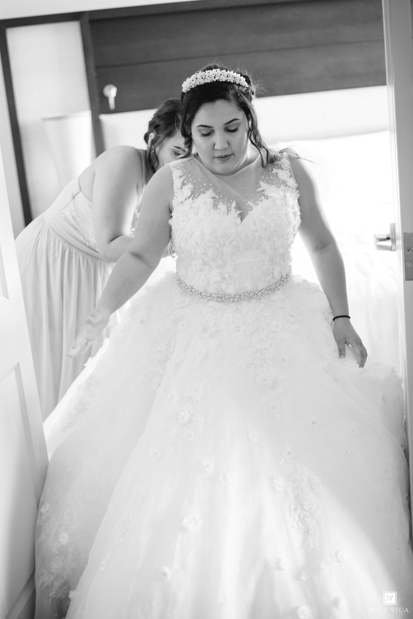 Bride in a floral lace ball gown with crystal belt and tiara, assisted by bridesmaid in black and white photo.