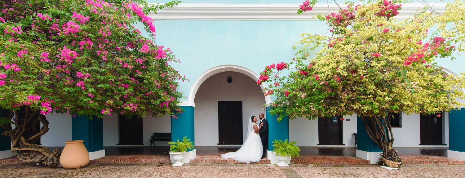 A couple shares a romantic kiss at a colorful colonial venue with bougainvillea and turquoise arches on their wedding day.