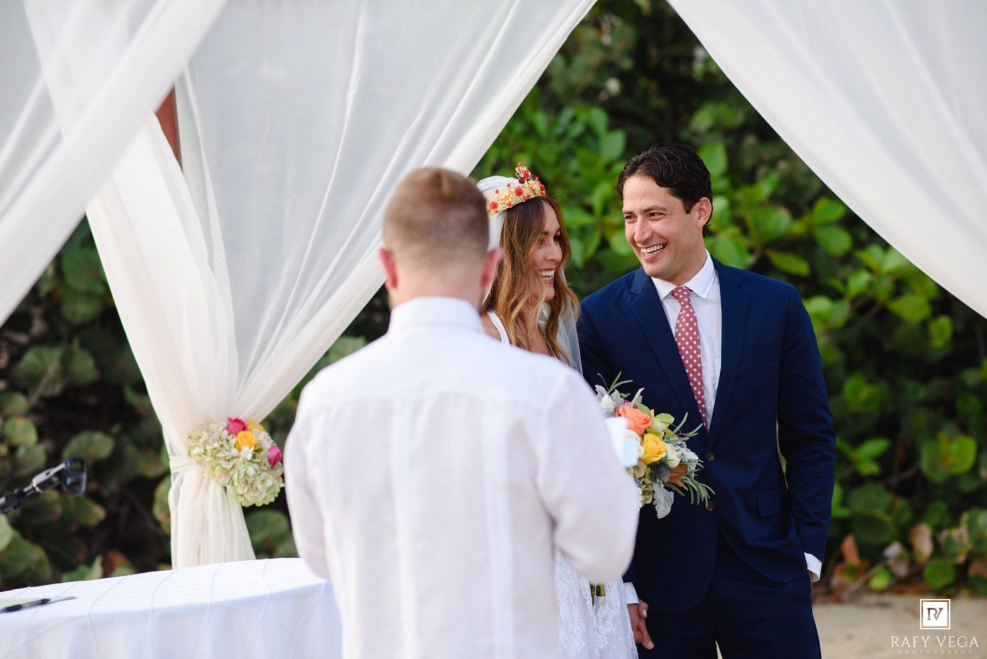 Outdoor wedding ceremony with bride in floral crown, groom in navy suit, and officiant under white draped canopy.