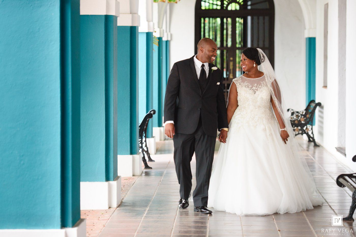 A bride and groom walk hand in hand along a elegant teal-columned hallway on their wedding day.