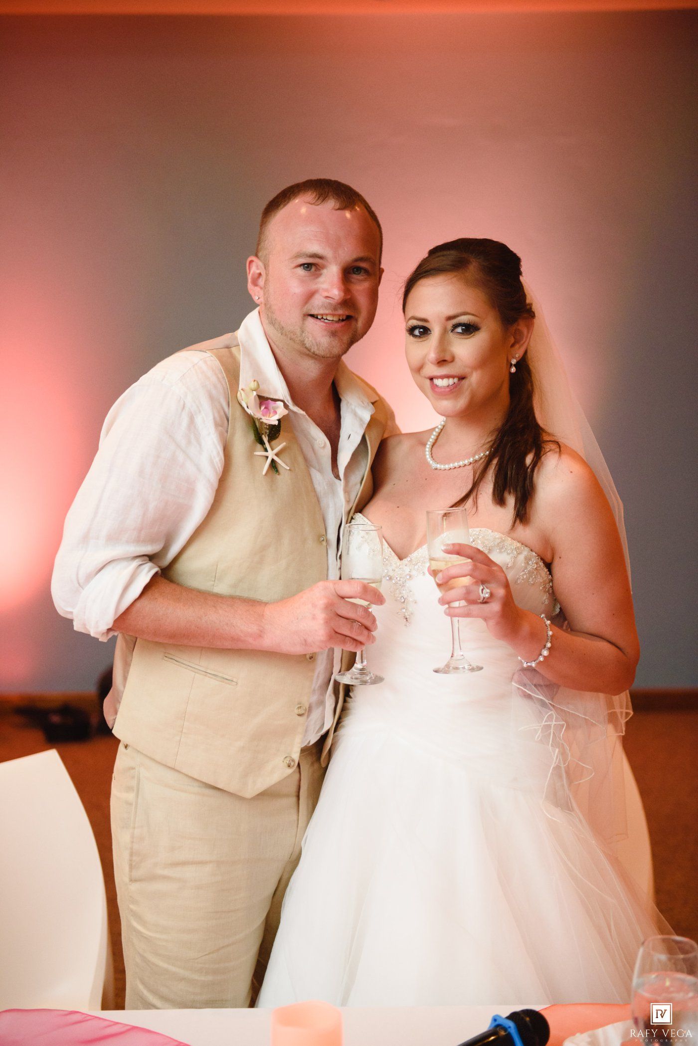 Bride and groom smiling and holding champagne glasses at their wedding reception, dressed in white gown and khaki suit.