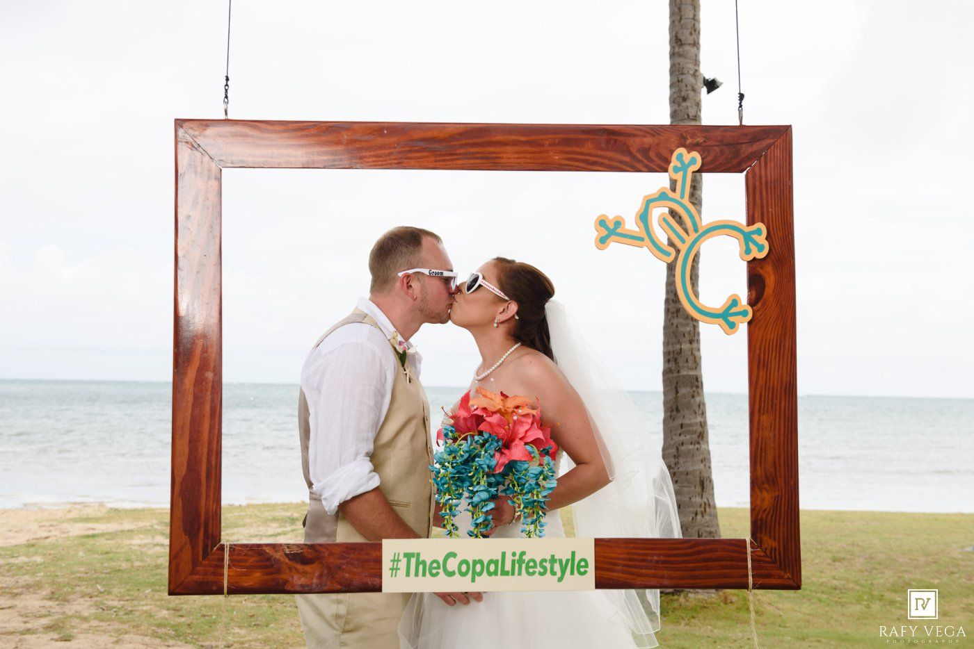 Bride and groom kissing inside a wooden frame photo prop on a tropical beach with #TheCopaLifestyle hashtag.