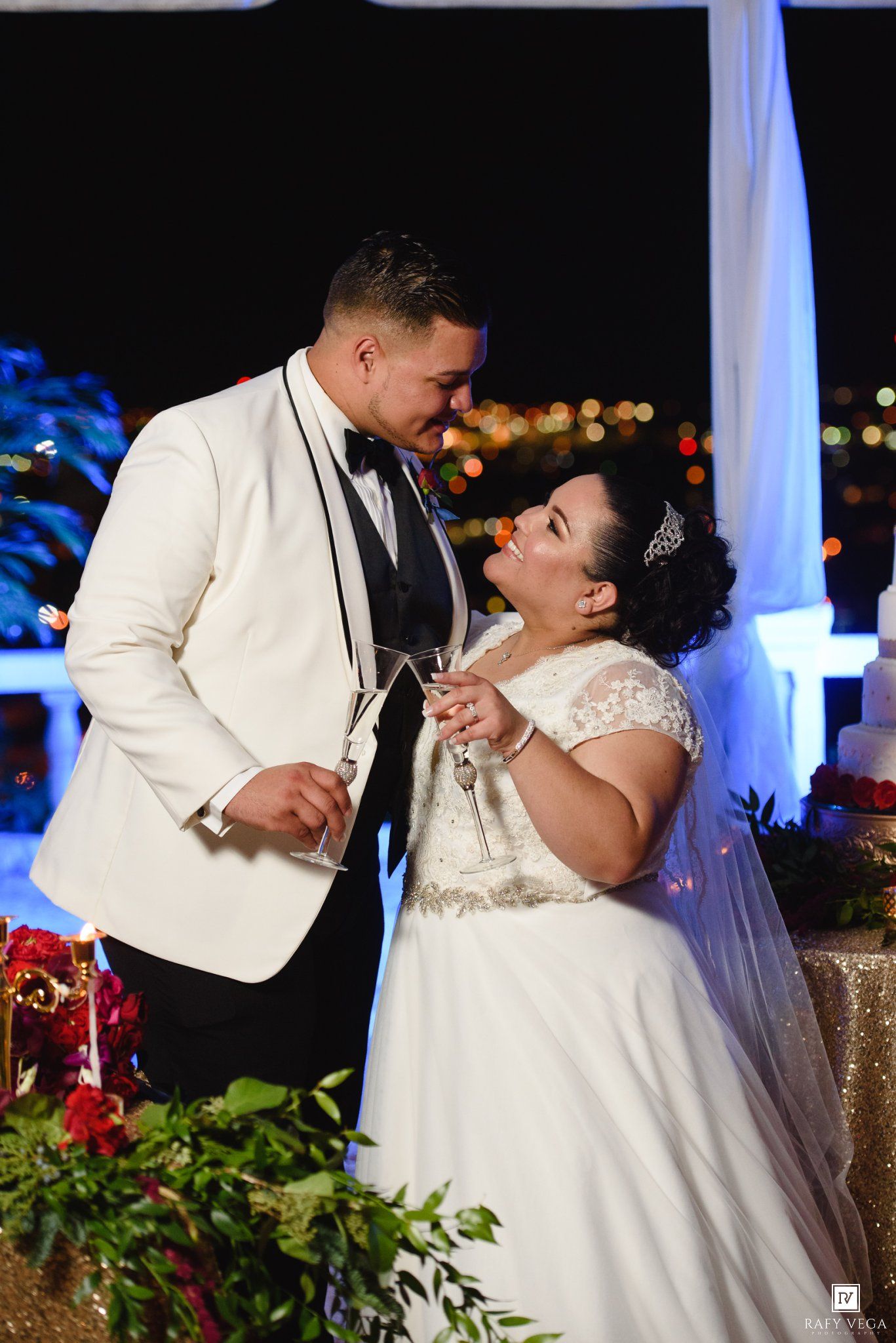Newlyweds share intimate toast with champagne flutes near wedding cake at romantic outdoor night reception.