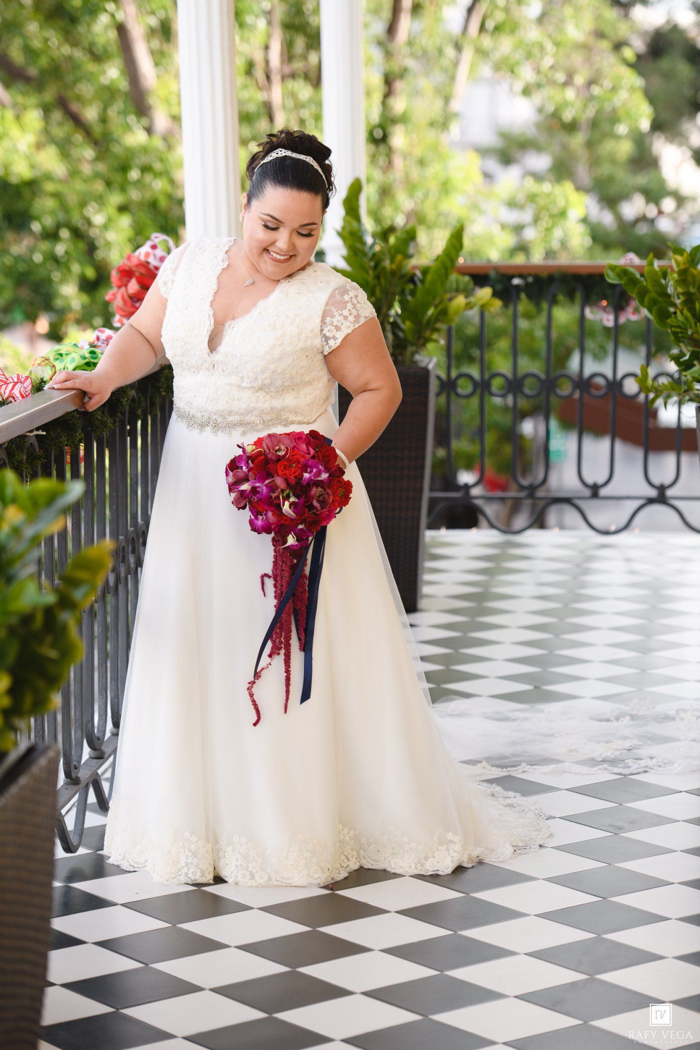 Bride in white lace A-line gown holding red bouquet on checkered balcony.