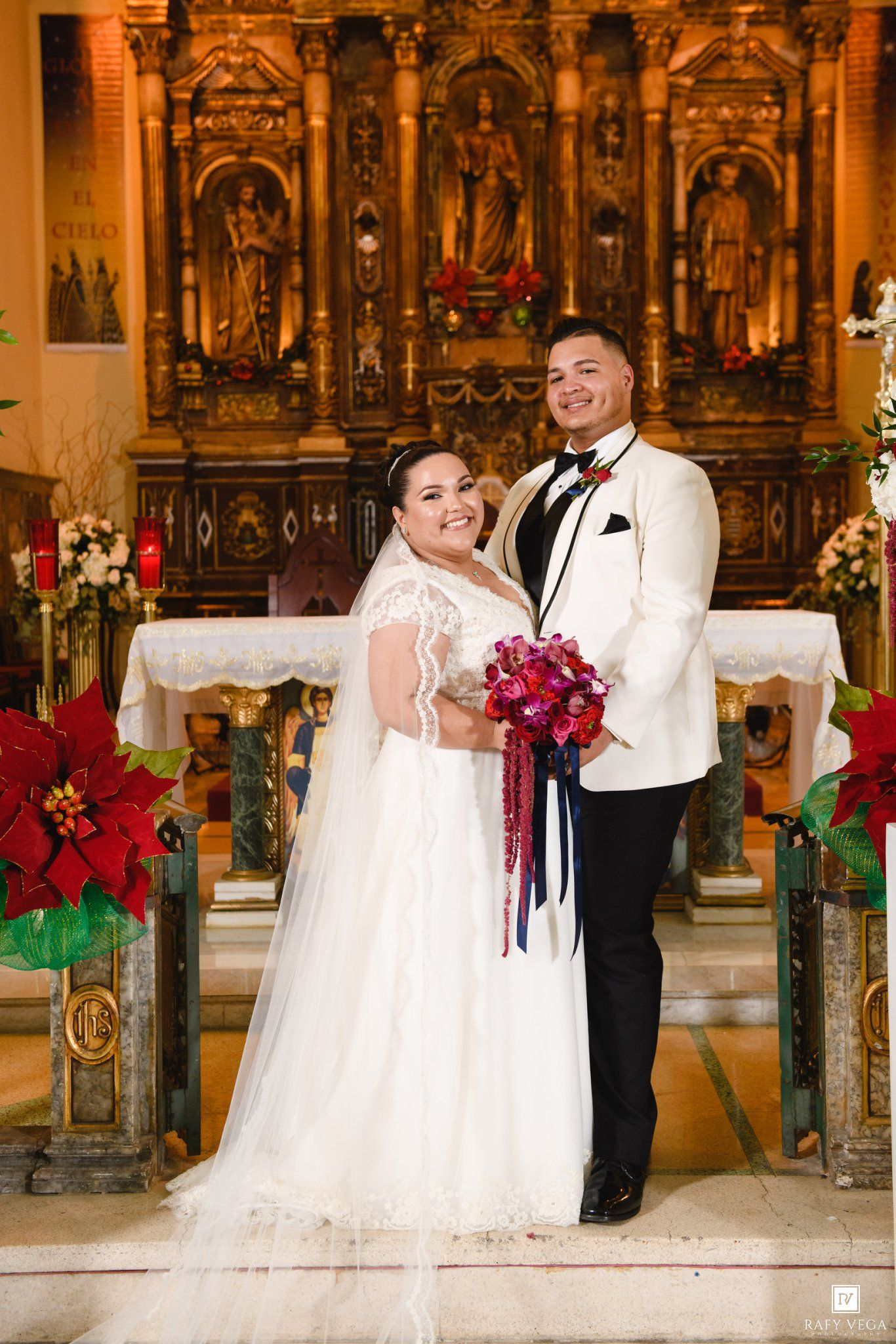 Newlyweds pose together at ornate church altar, bride holding red bouquet in wedding portrait.