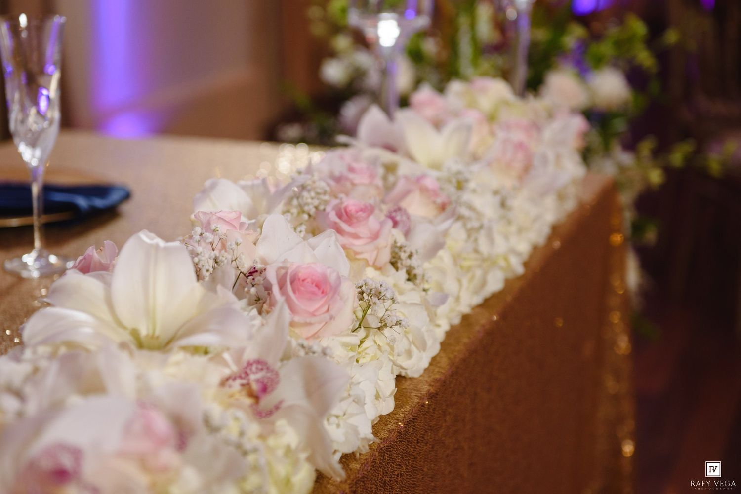 Elegant wedding table centerpiece featuring pink roses, white lilies, and hydrangeas on a gold glitter tablecloth.
