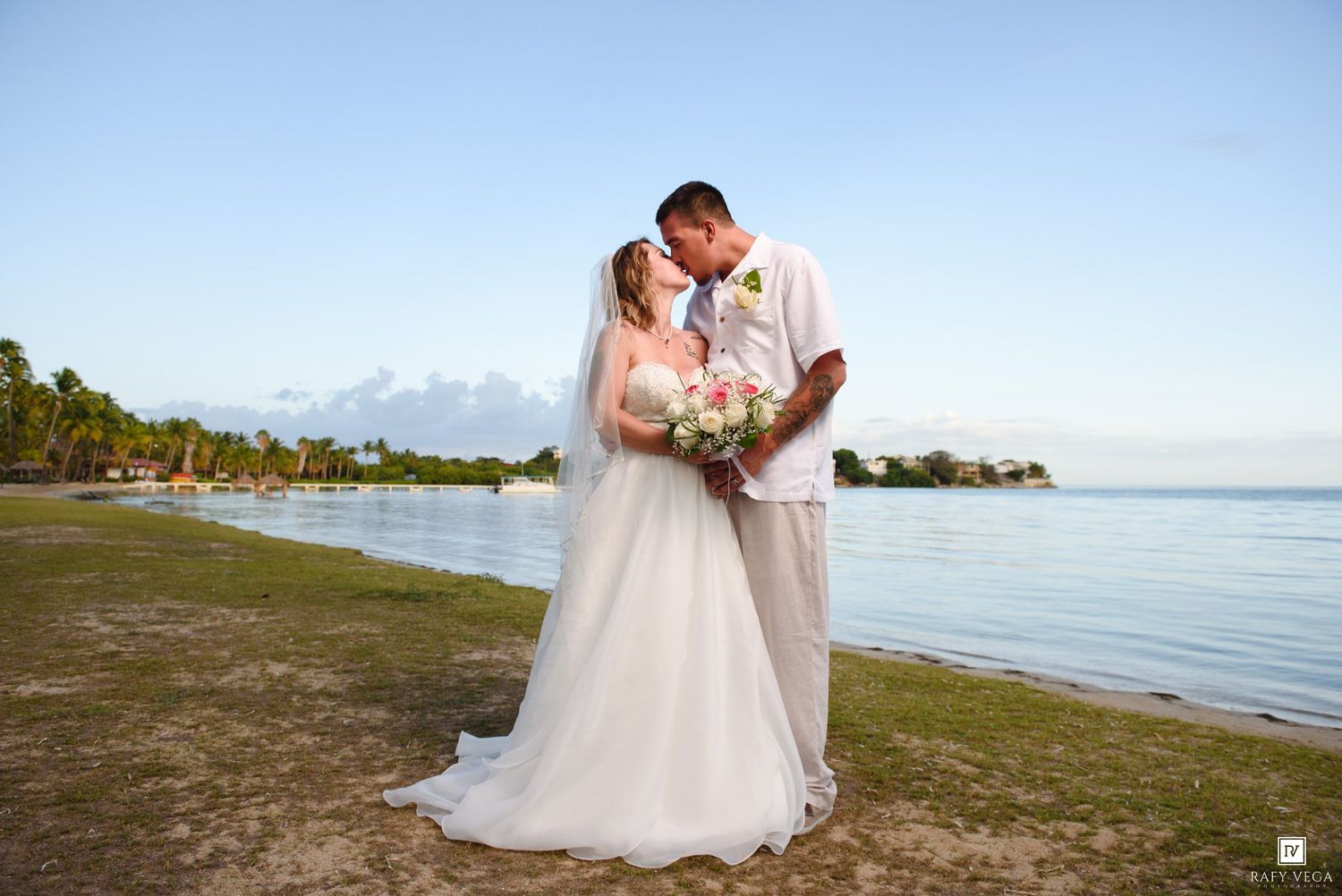 Bride and groom share a kiss on a tropical beach at sunset, bride holding a pink and white bouquet.