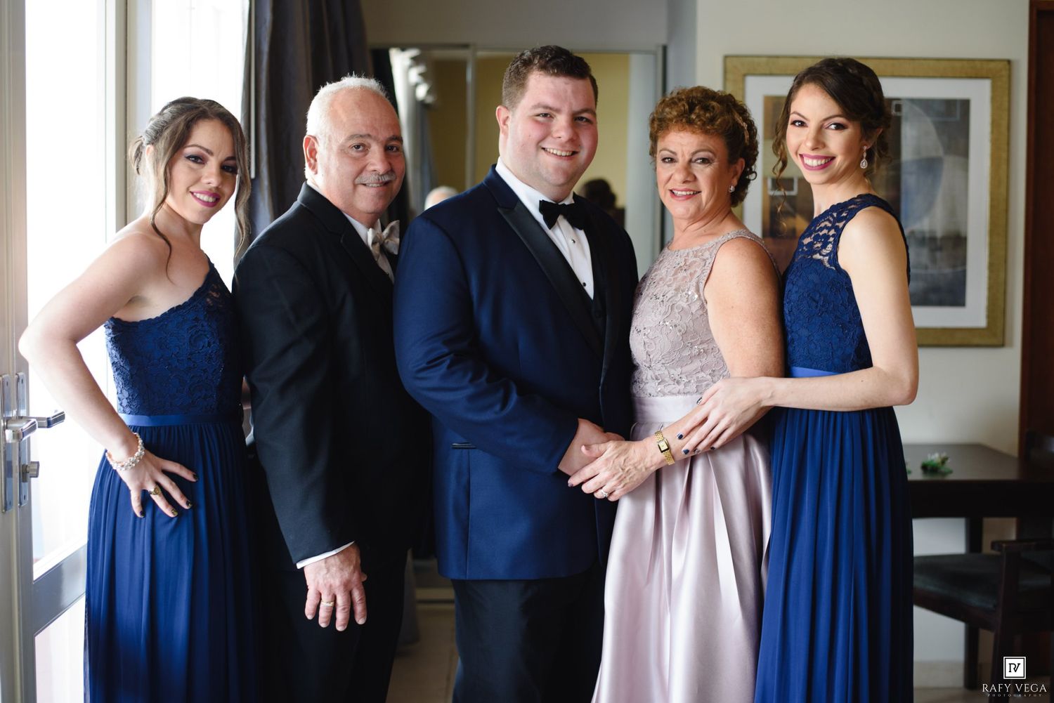 Family group photo in formal attire, two women in blue gowns flanking three people in elegant wedding party dress.