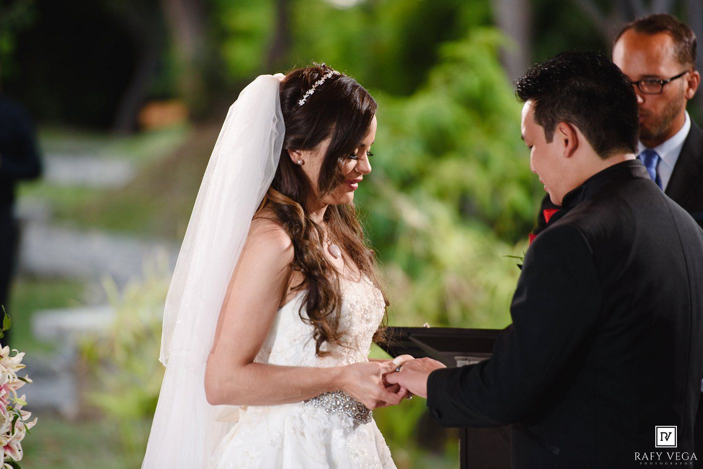 Bride smiling while exchanging rings with groom during outdoor wedding ceremony, officiant standing behind them.