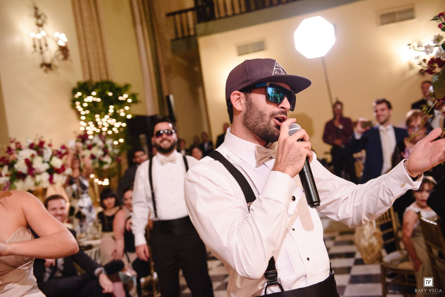 Man in cap and suspenders singing into microphone at elegant wedding reception with floral decorations.