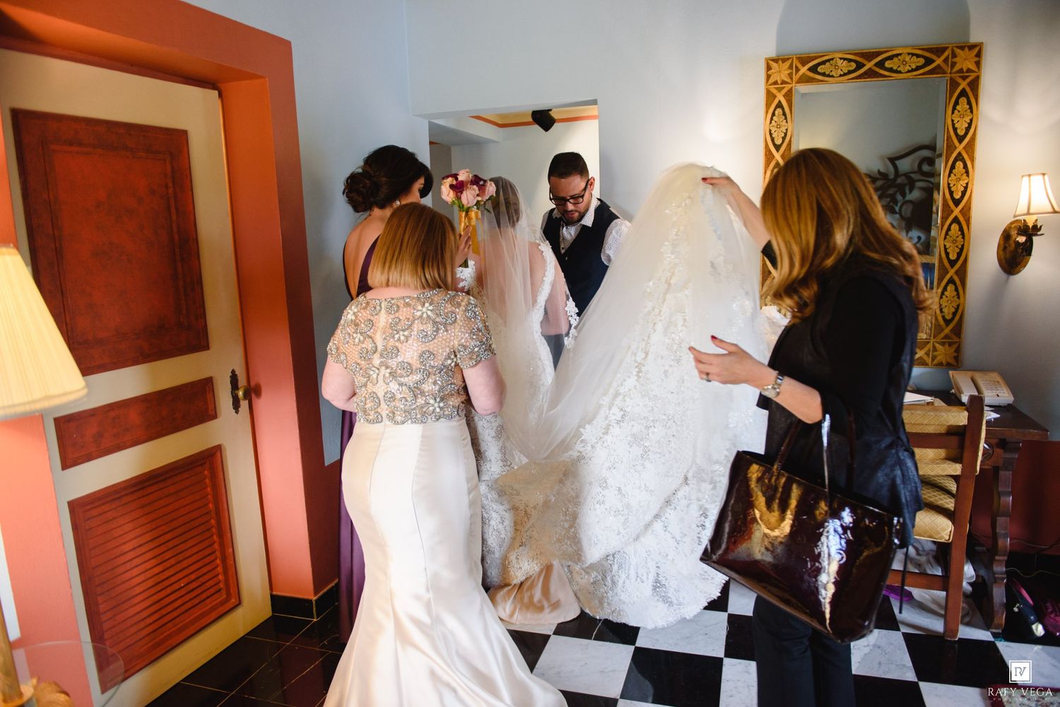 Bride in white gown with veil being assisted by wedding guests in elegant room with checkered floor.