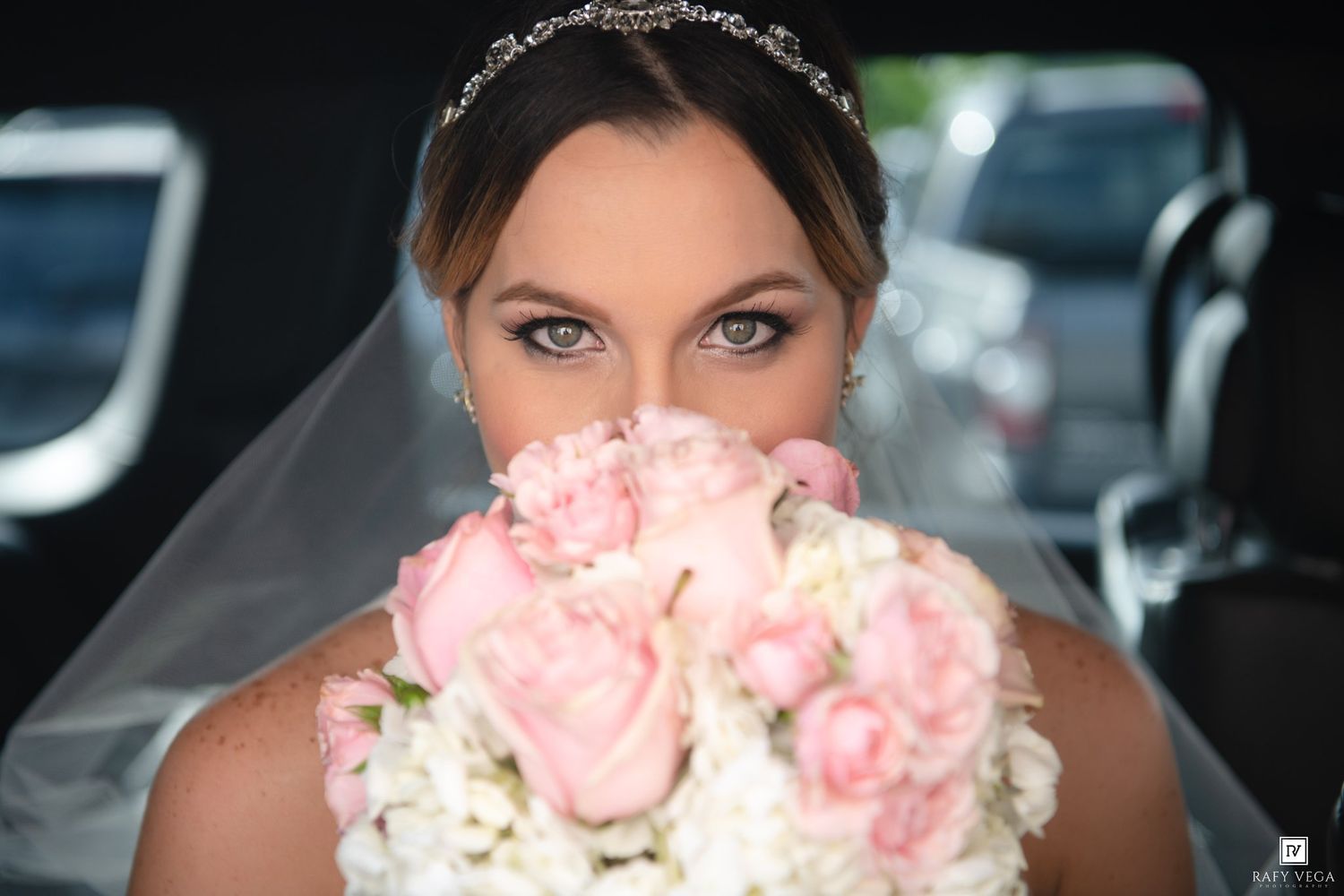 Bride in wedding veil holds pink rose bouquet up to her face, showcasing stunning blue eyes and tiara.