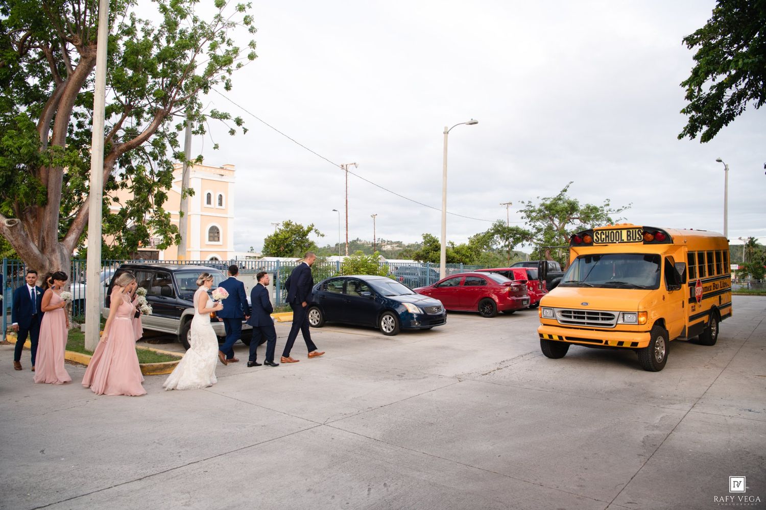 Wedding party in formal attire gathered outside near a yellow school bus and parked cars on a sunny day.