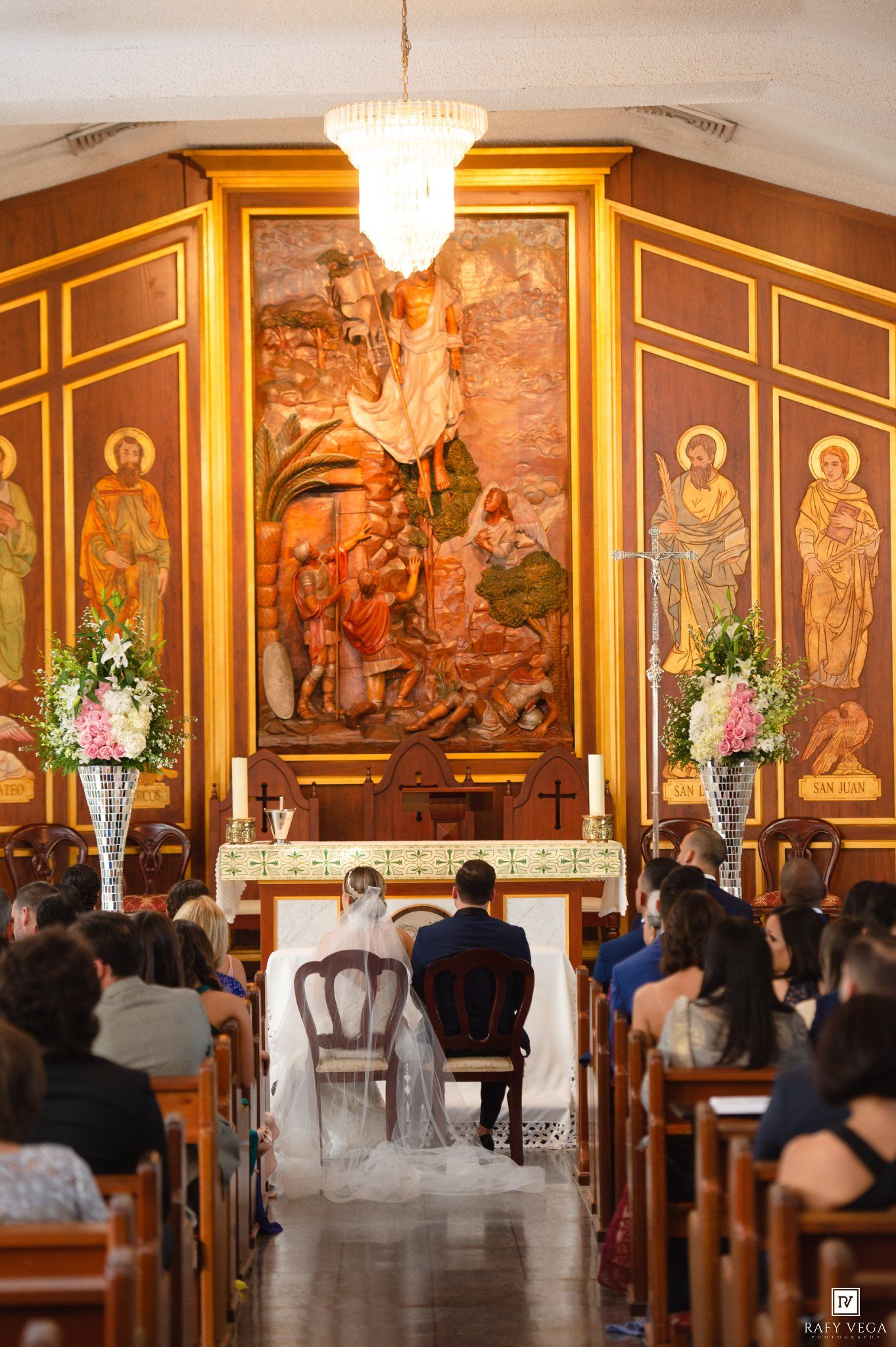 Bride and groom kneeling at church altar during wedding ceremony with ornate religious murals and floral arrangements.