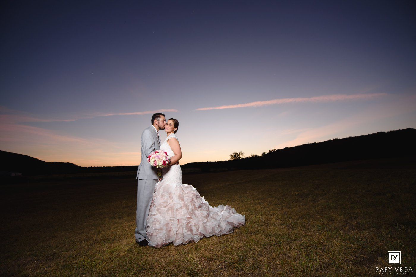 Bride and groom share a romantic kiss at sunset on a green field, with a stunning pink and blue sky backdrop.