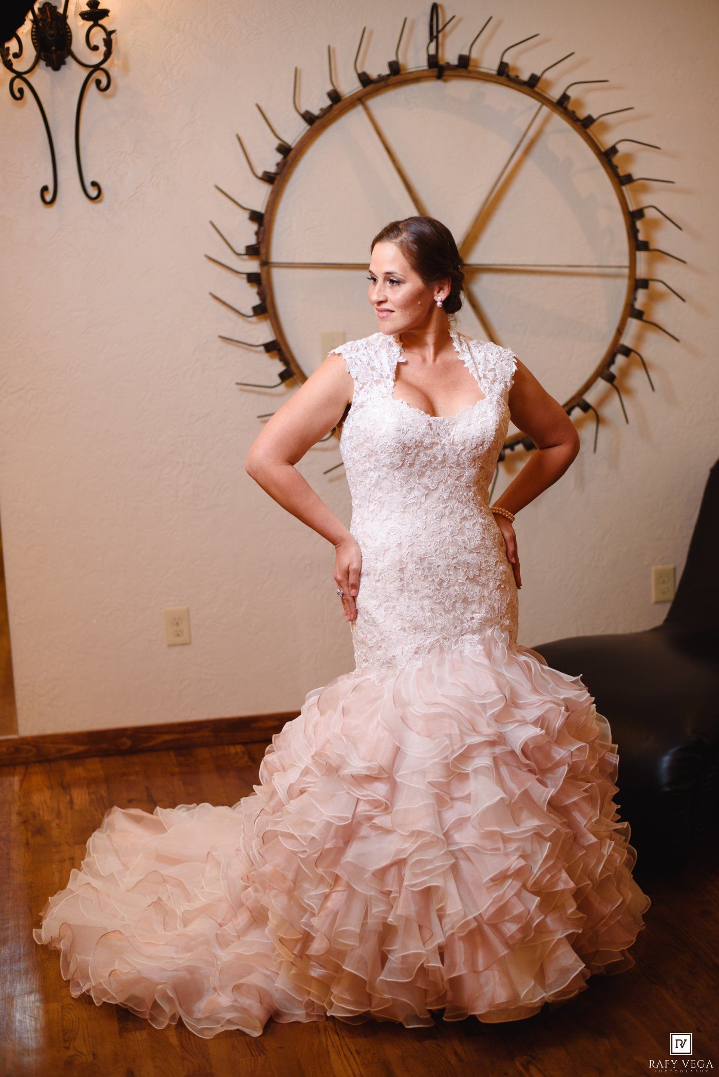 Bride in a mermaid-style lace and ruffle wedding gown poses elegantly in front of a decorative wagon wheel wall art.