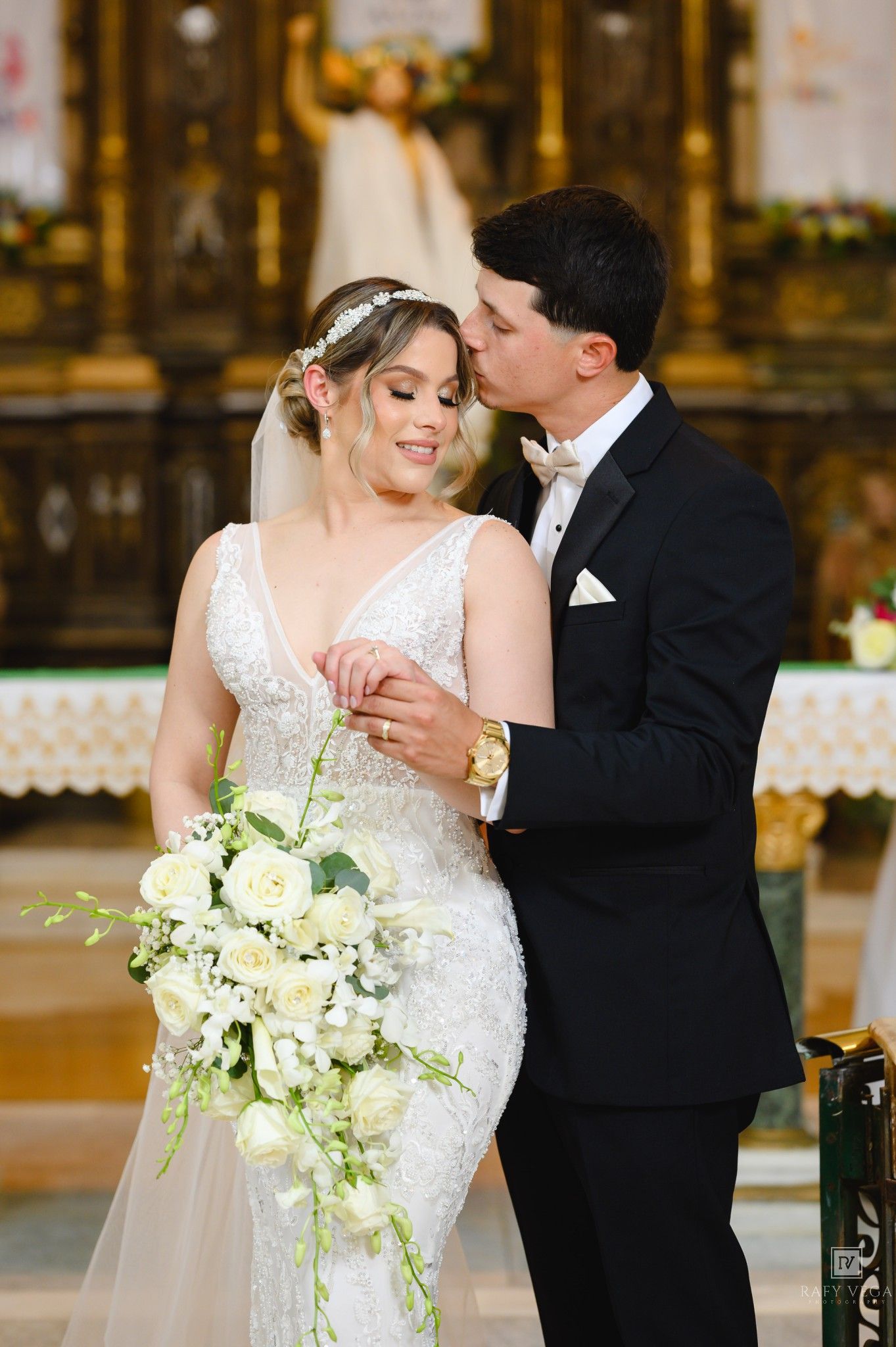 Bride and groom share a tender moment at the church altar, she holds a white cascading bouquet in a beaded gown.