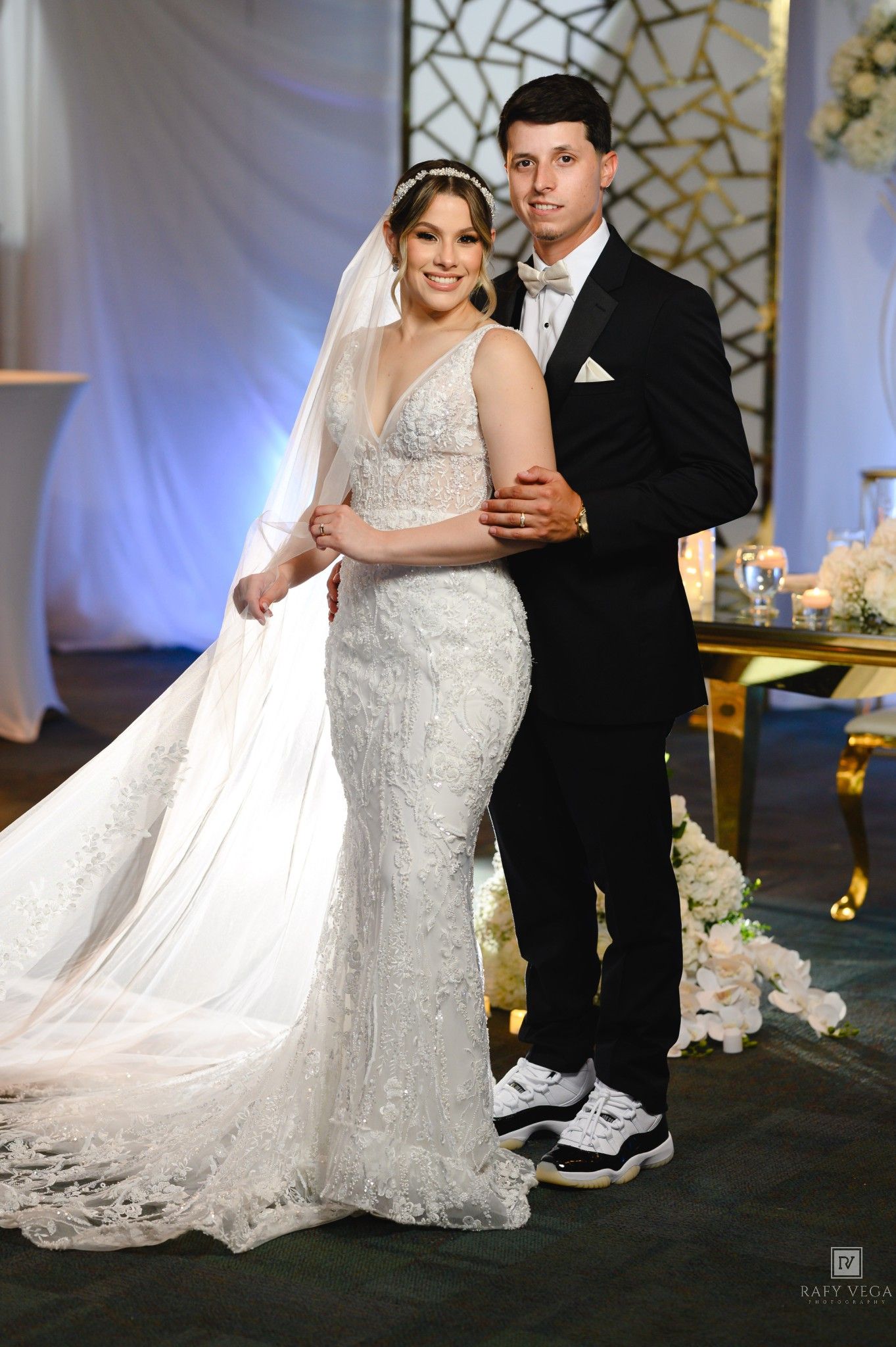 Bride in lace mermaid gown with veil and groom in black tuxedo pose together at elegant wedding reception.