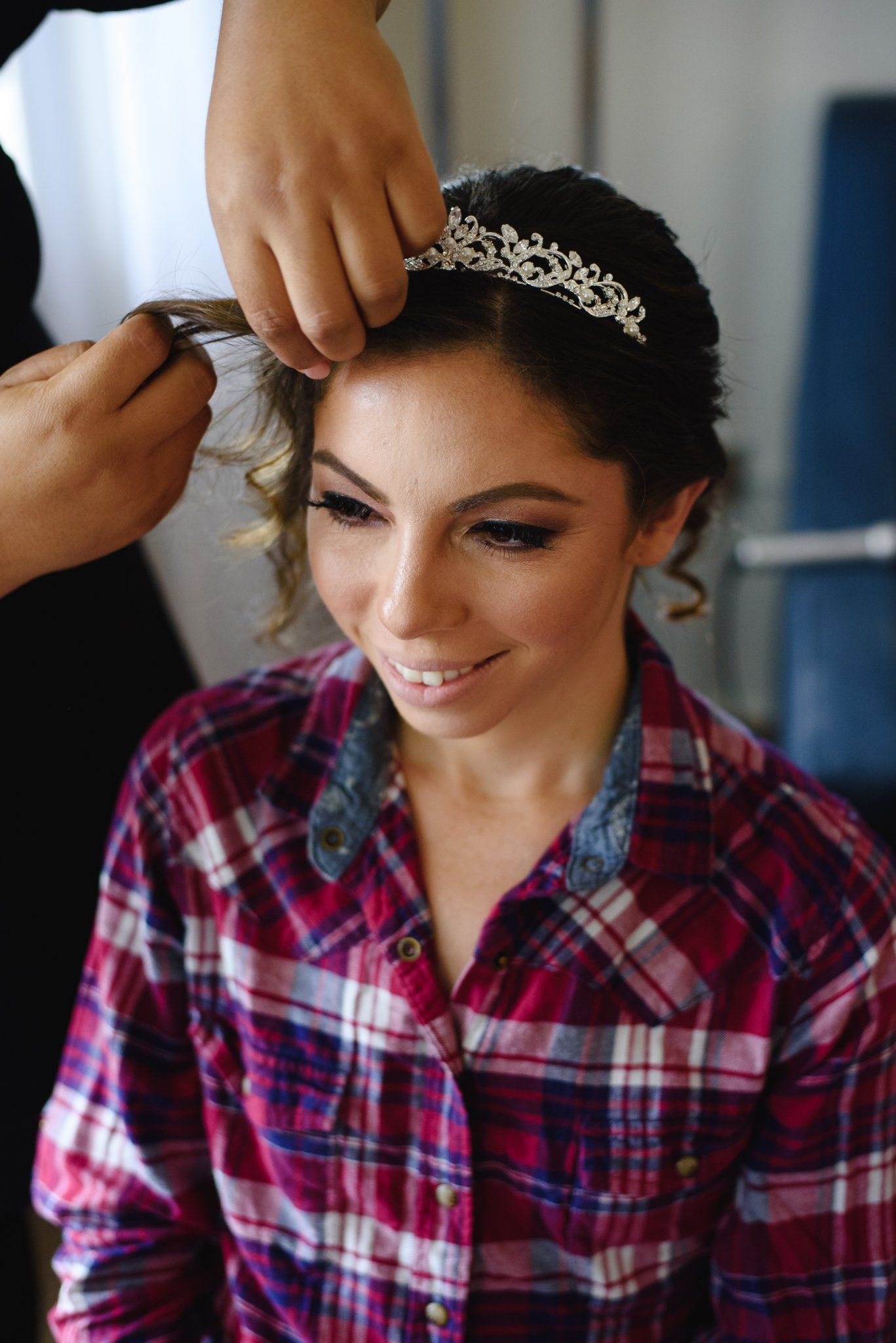 A smiling bride wearing a tiara and plaid shirt gets her hair styled by a hairstylist during wedding preparations.