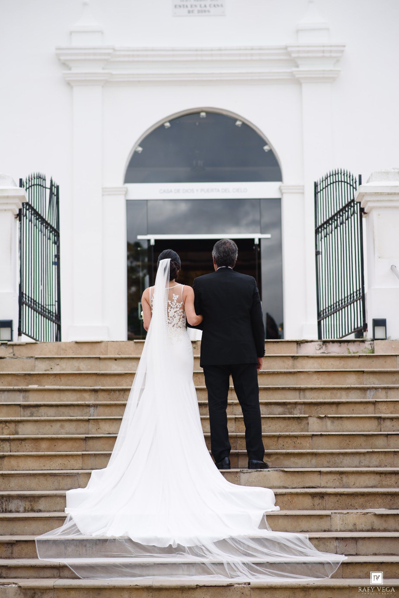Bride and groom stand on church steps, her lace gown and long veil trailing behind, viewed from behind.