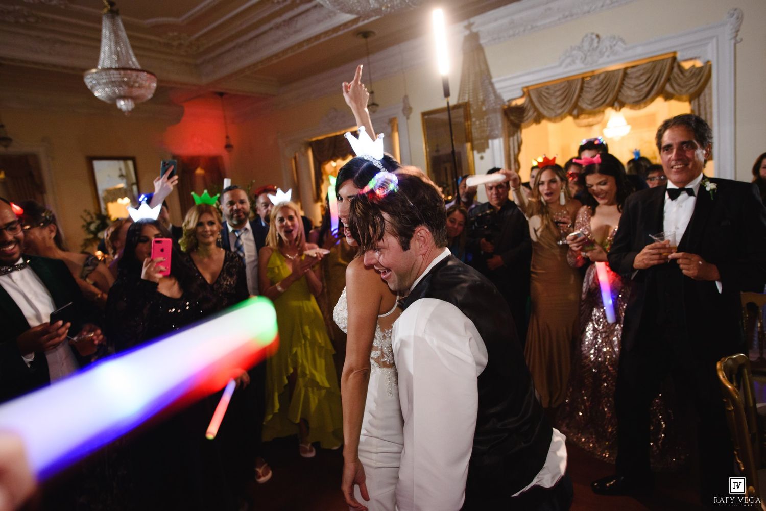 Bride and groom share a kiss surrounded by guests with colorful glow sticks at wedding reception.