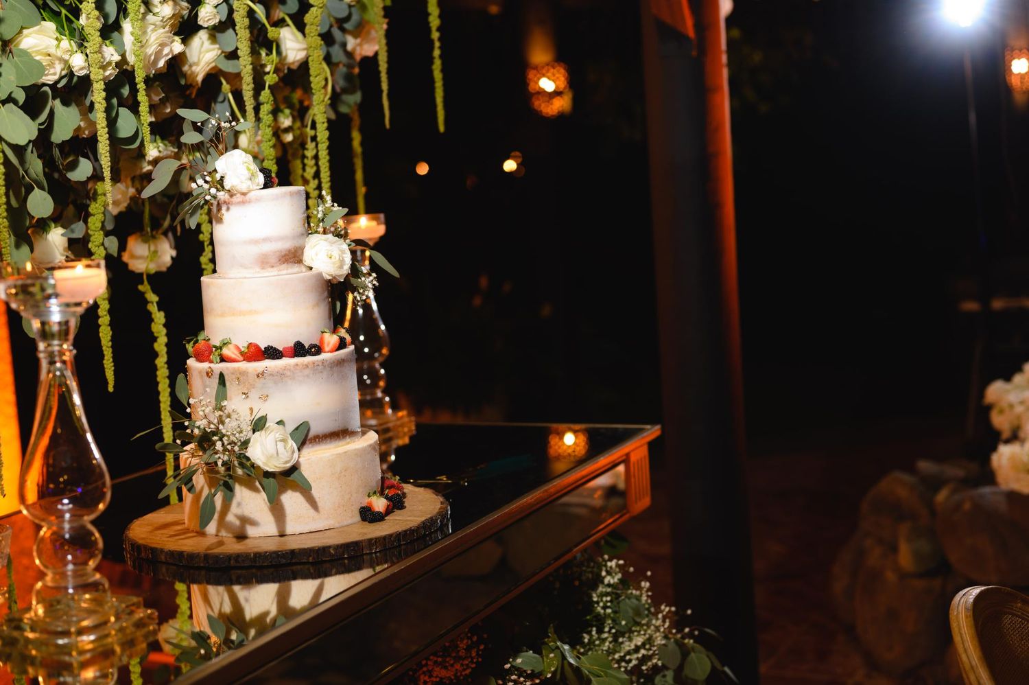 Elegant three-tier white wedding cake adorned with flowers on a wooden stand, beautifully lit at an outdoor night reception.
