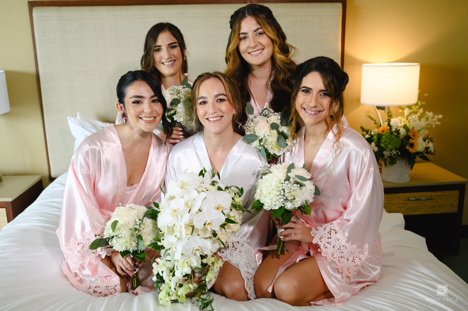 Five bridesmaids in pink robes sitting on a hotel bed holding white floral bouquets on a wedding day.
