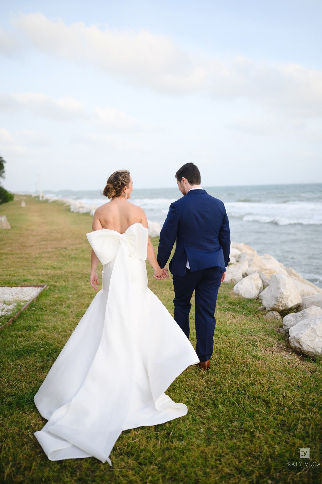 Bride and groom walk hand-in-hand along a scenic oceanside path, bride wearing a stunning white gown with large bow.