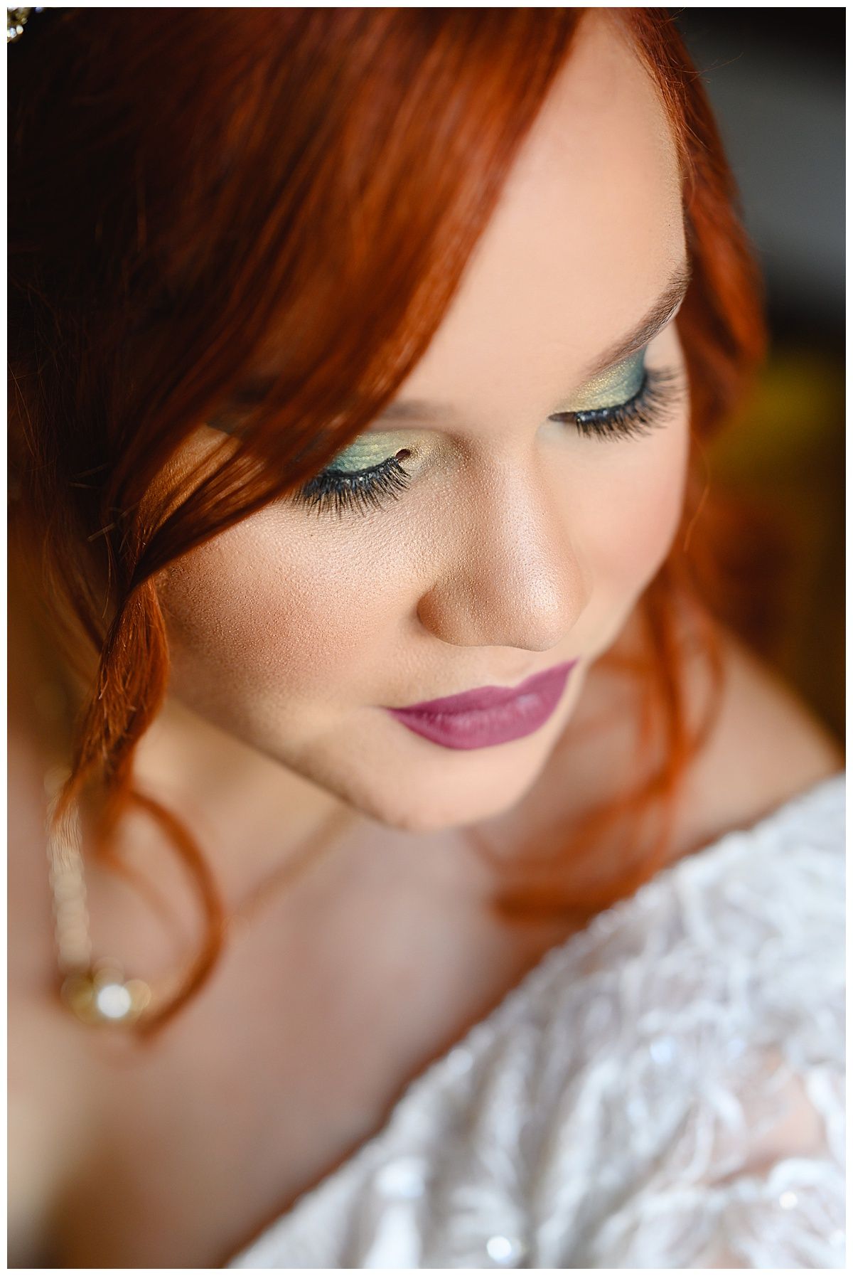 Close-up portrait of a red-haired bride with green eyeshadow, long lashes, and berry lips in a white lace dress.