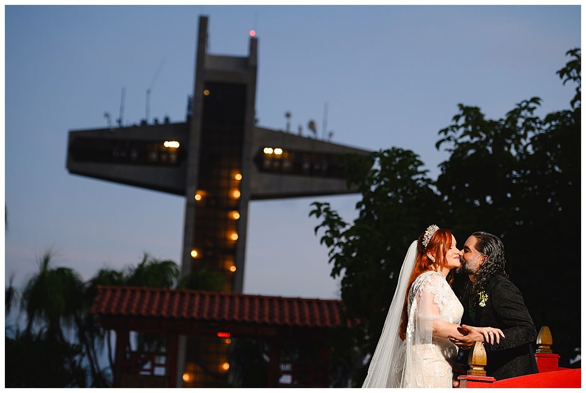Bride and groom share a romantic kiss at dusk with an illuminated cross and tropical trees in the background.