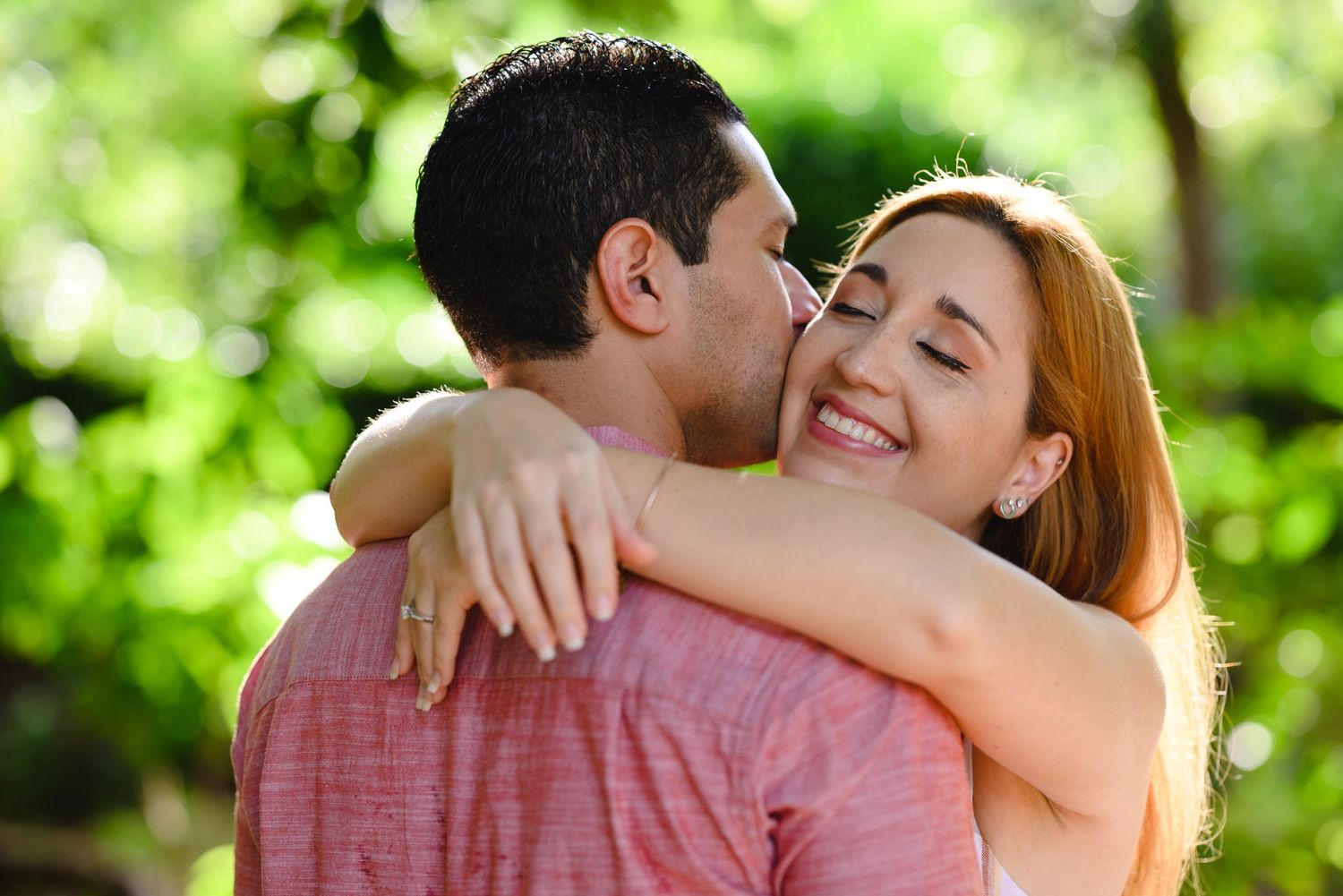 A happy couple embraces outdoors, woman smiling brightly as man kisses her cheek in a lush green setting.