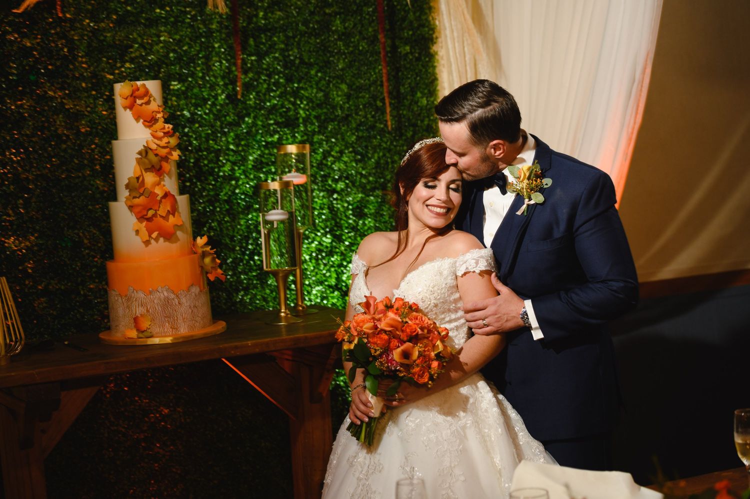Bride and groom share a romantic moment beside an orange floral wedding cake against a lush green hedge backdrop.