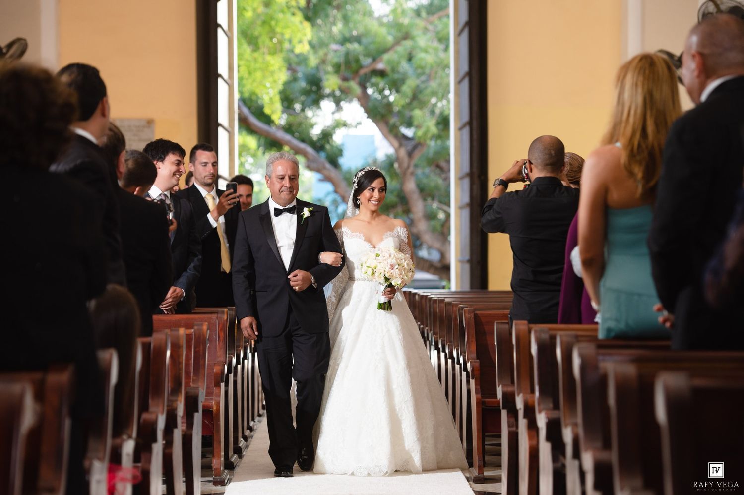 Father escorting bride down the church aisle surrounded by wedding guests during ceremony.