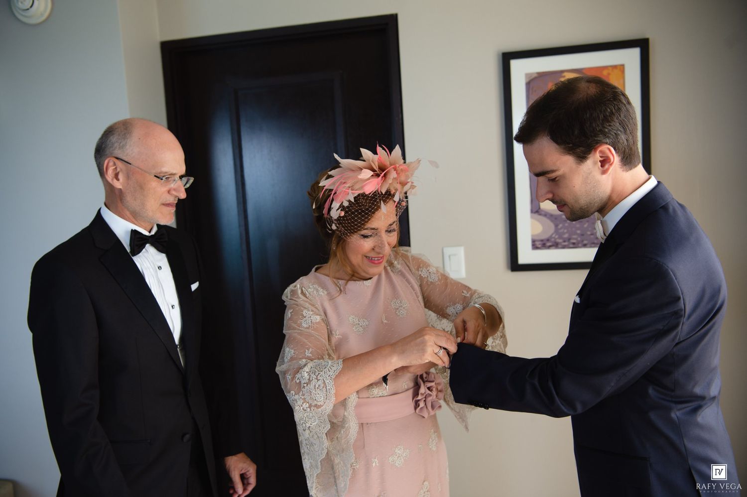 Elegant wedding preparation scene with bride in pink dress and floral headpiece assisted by two men in formal attire.