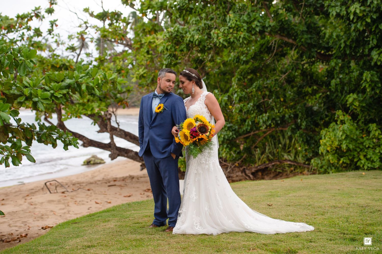 Bride in white gown holding sunflower bouquet stands with groom in blue suit on tropical beach lawn.
