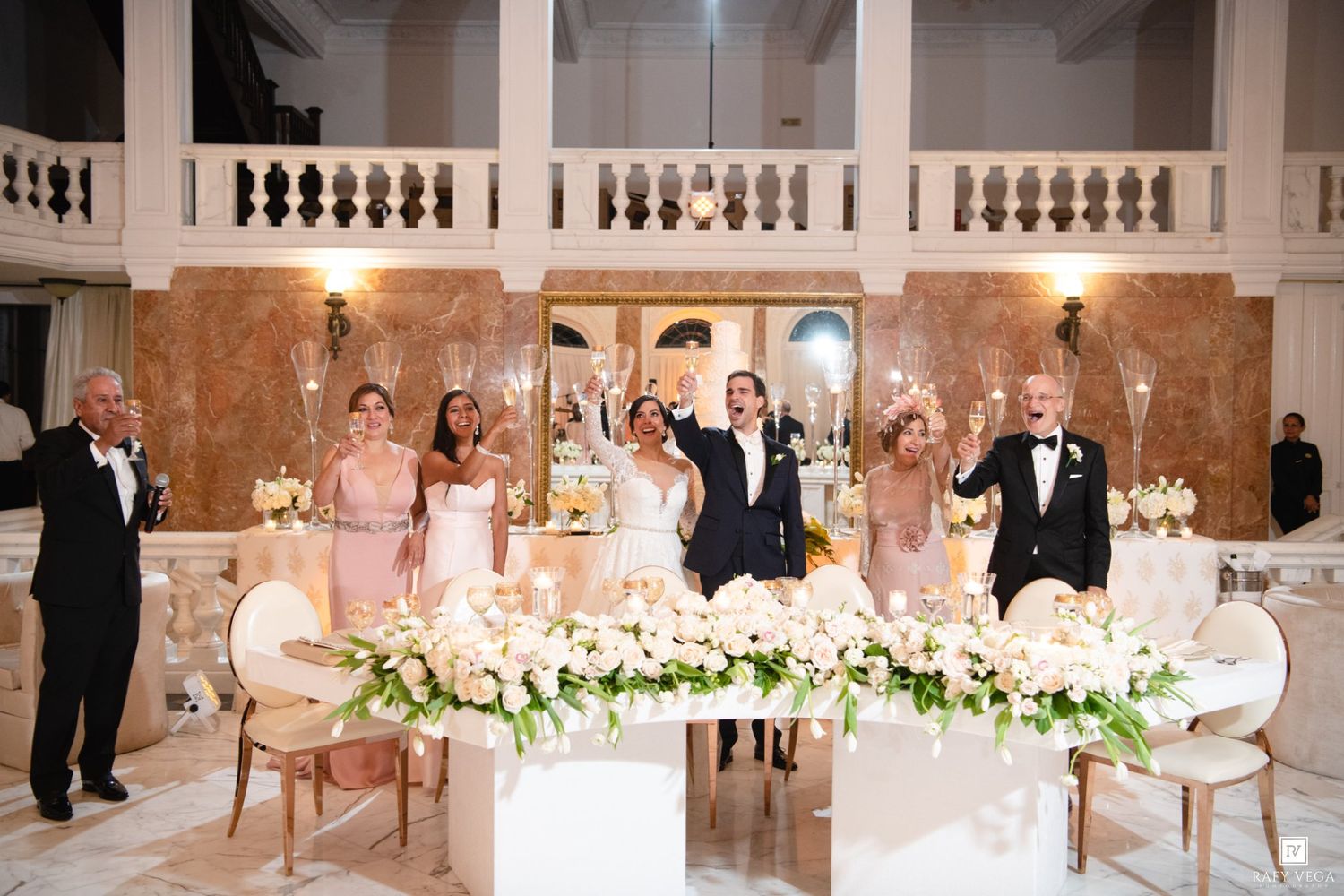 Wedding party toasts at elegant reception with white floral arrangements, bridesmaids in pink, grand marble venue.