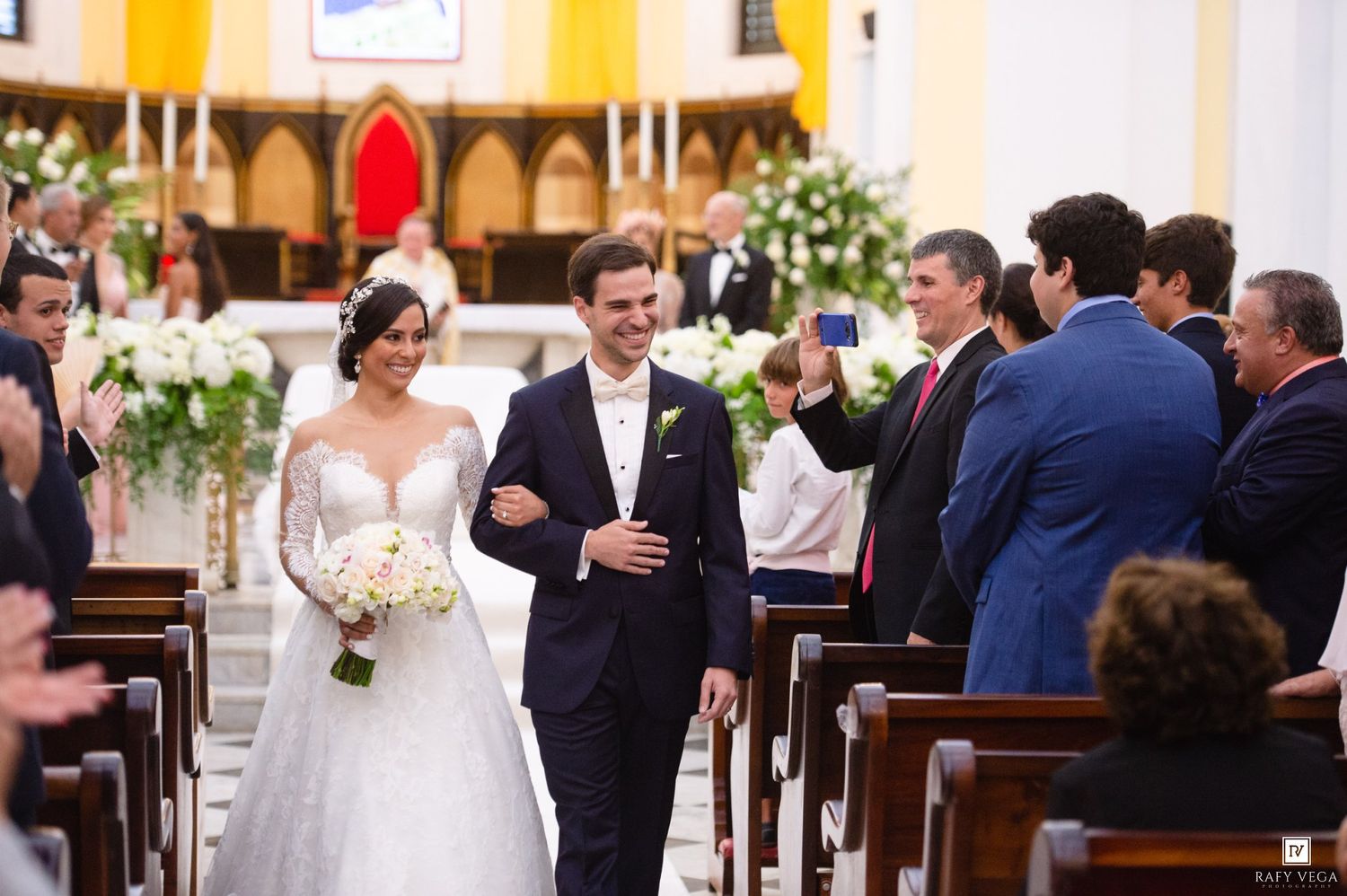 Bride and groom walk down church aisle after wedding ceremony, surrounded by cheering guests in elegant venue.