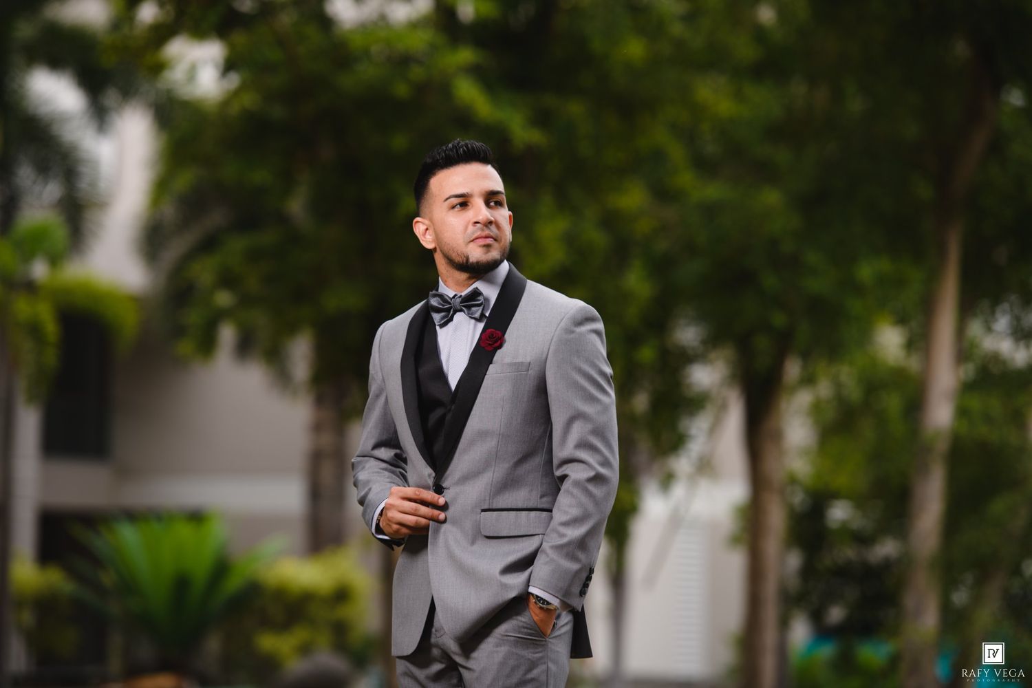 Groom in stylish grey three-piece suit with black bow tie posing outdoors among green trees.