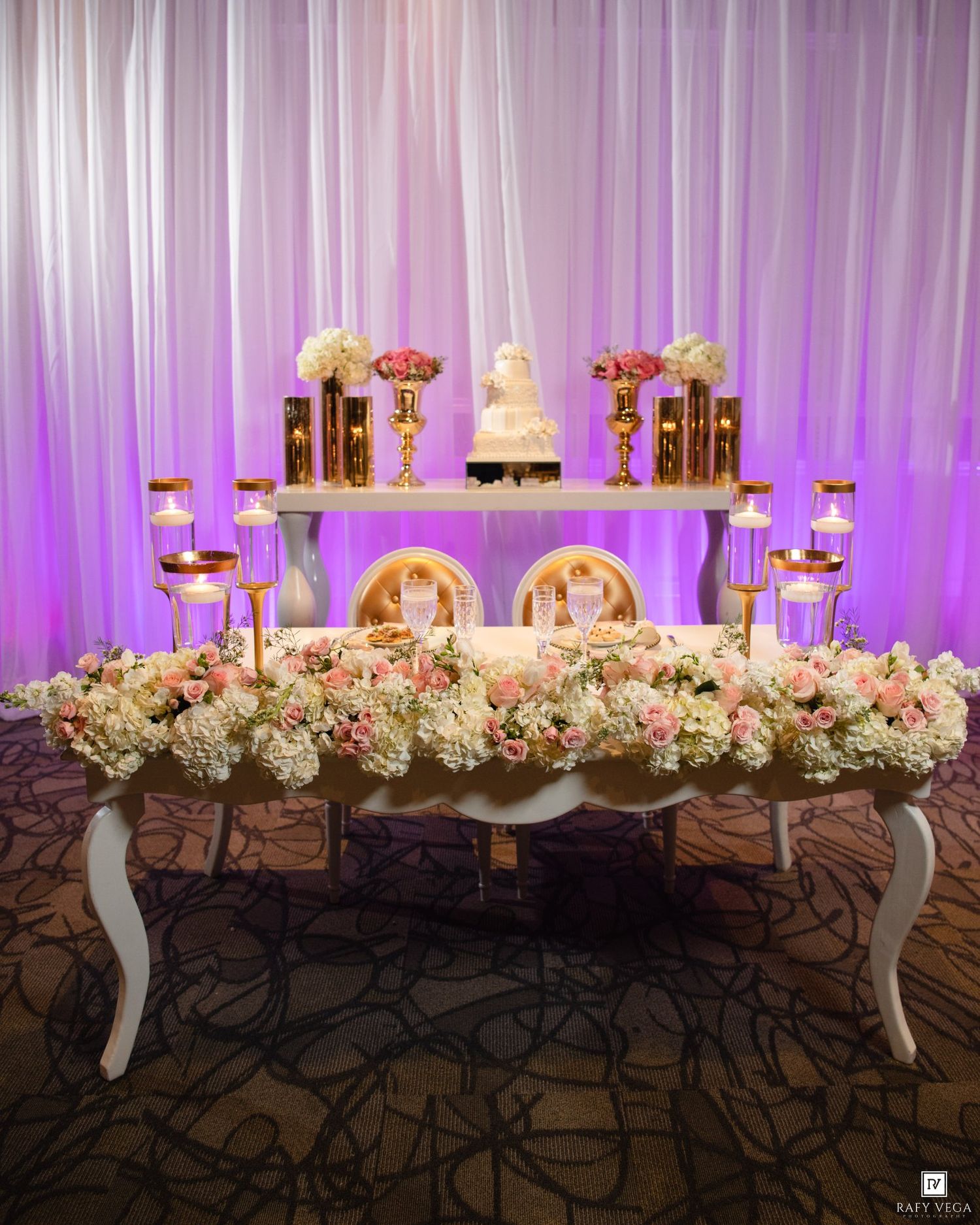 Elegant wedding sweetheart table adorned with pink roses, white florals, gold candelabras, and a tiered cake against purple draping.
