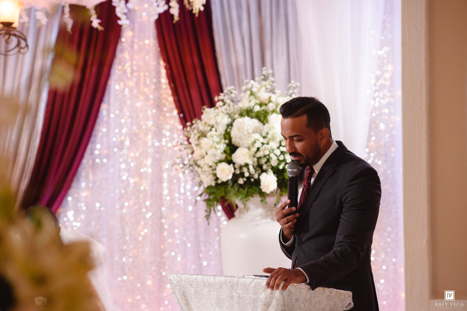 A man in a dark suit speaks into a microphone at a wedding reception podium with floral decor and twinkling lights.