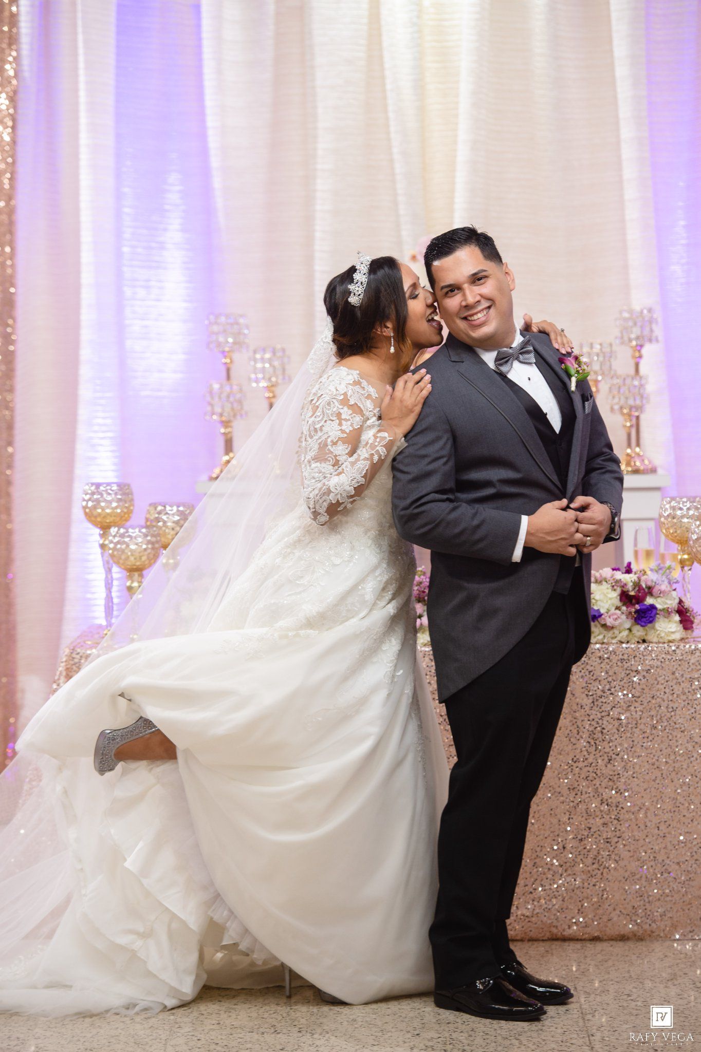 Bride in lace gown kisses groom in gray tuxedo at elegant wedding reception with gold candelabras.