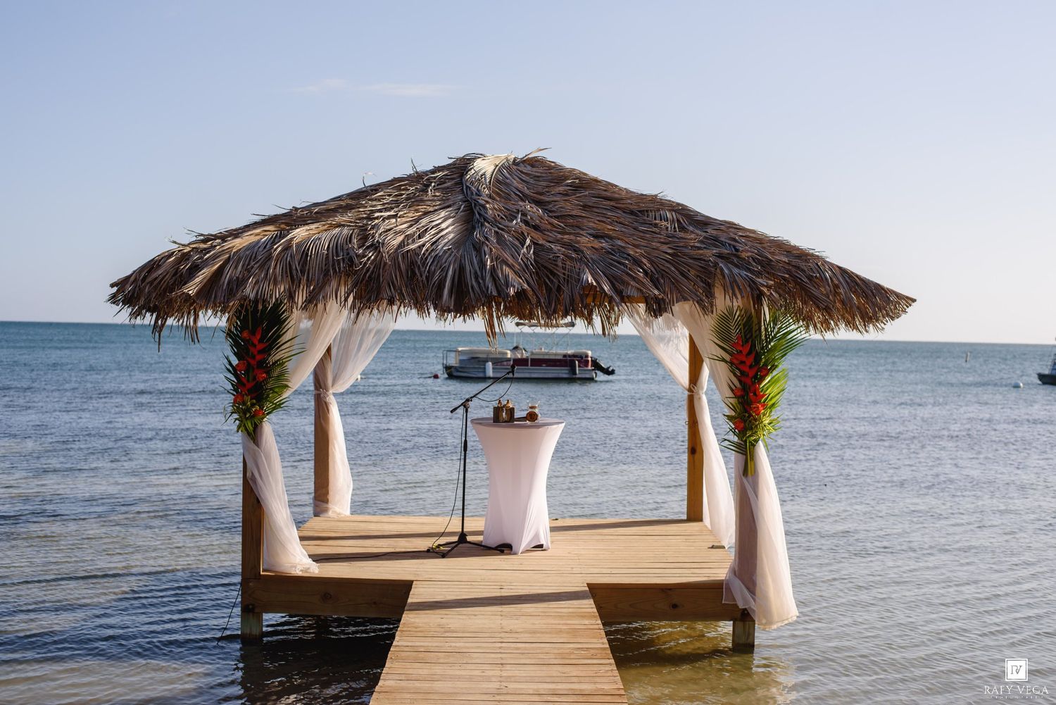 Tropical thatched gazebo on a wooden dock over turquoise Caribbean waters, decorated with white drapes for a beach wedding.