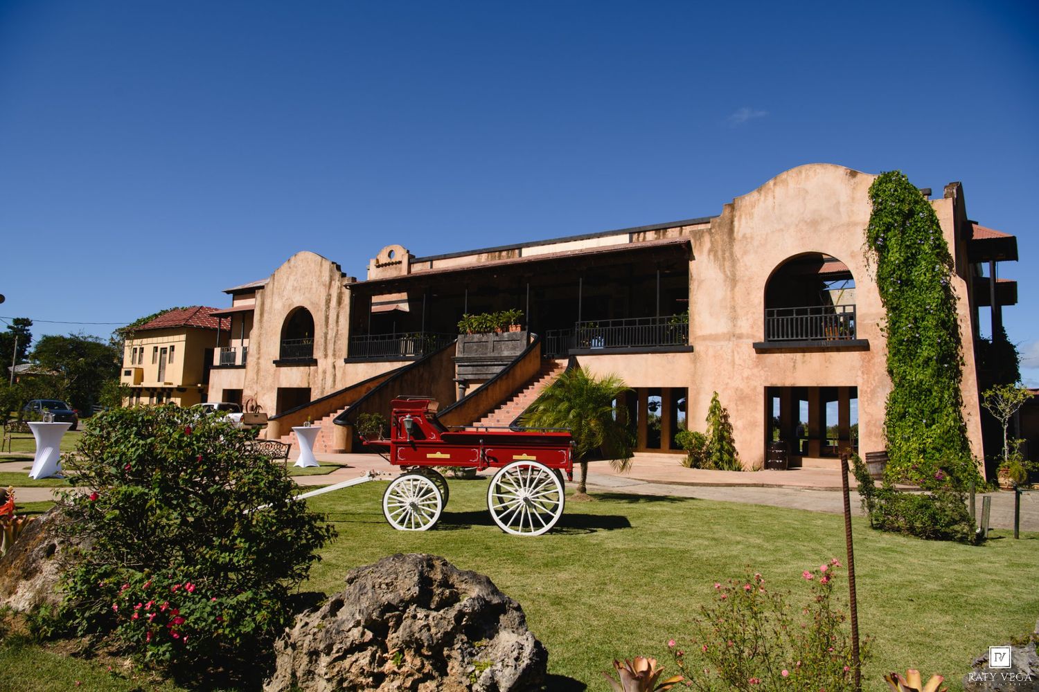Historic Spanish-style estate with red vintage horse carriage on lush green lawn under clear blue sky.
