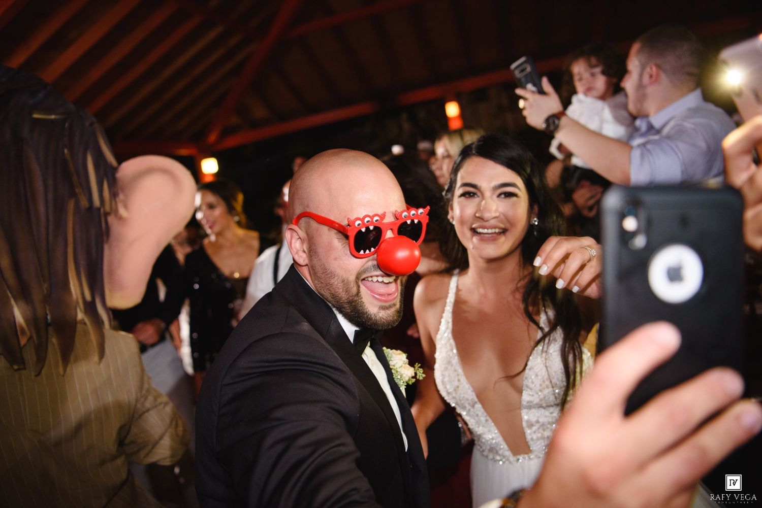 Bride and groom taking a selfie at wedding reception, groom wearing fun red clown glasses, guests celebrating in background.