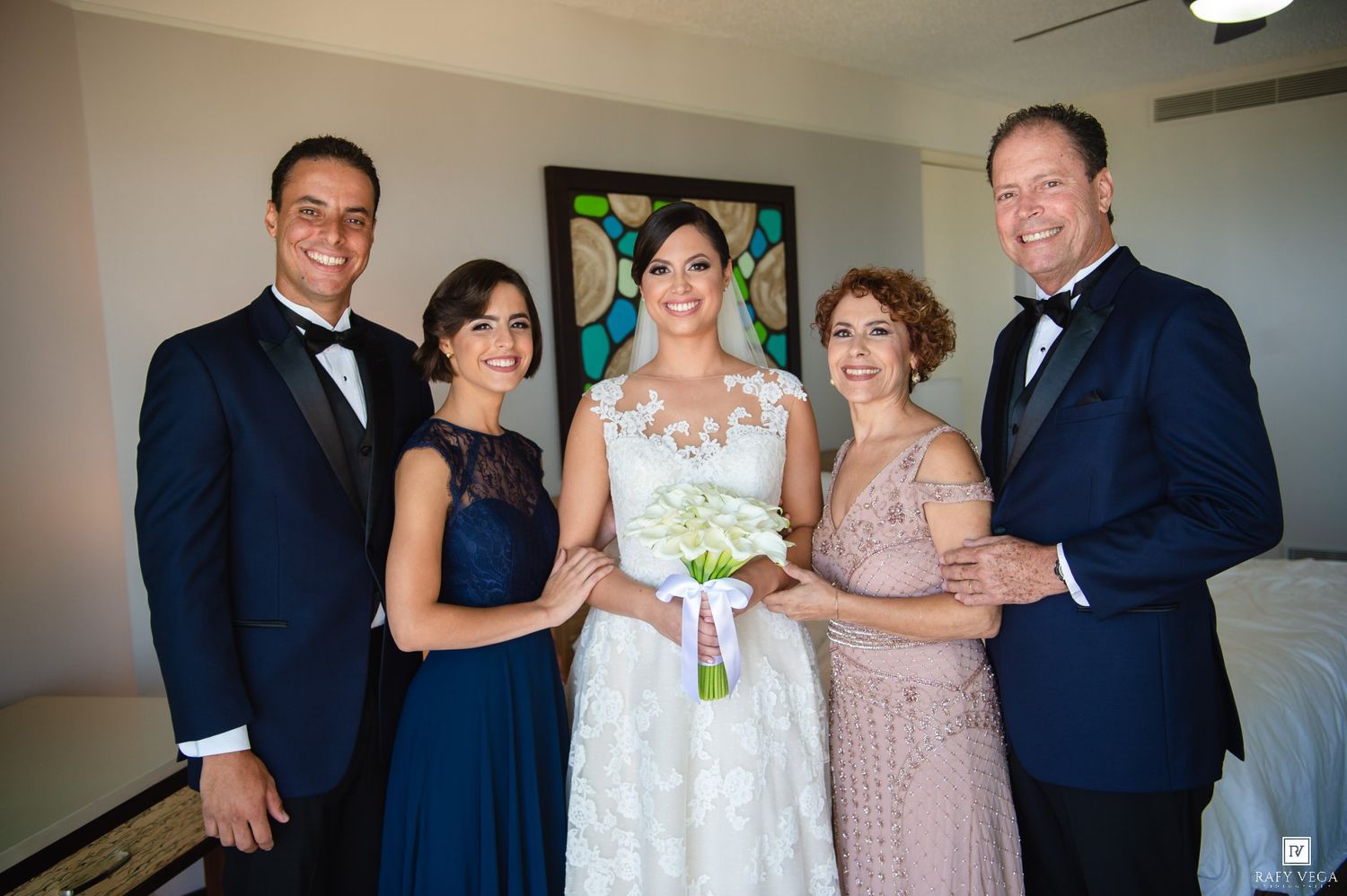 Bride in white lace gown holds bouquet surrounded by family in formal attire at wedding celebration.