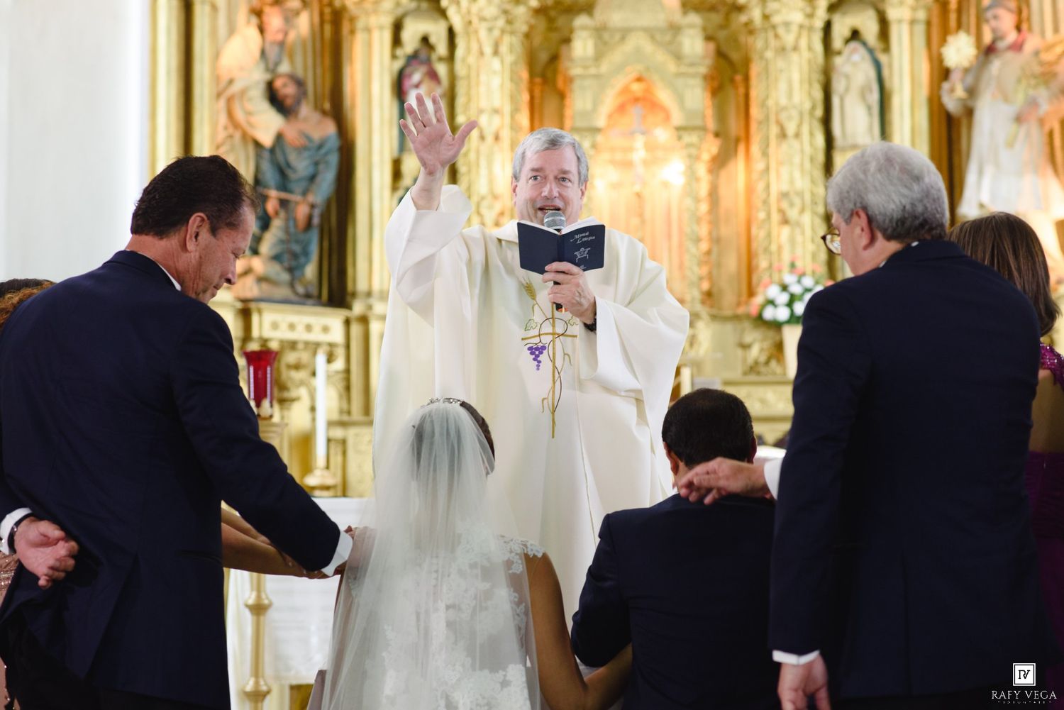 Priest blessing couple kneeling at altar during Catholic wedding ceremony in ornate church.