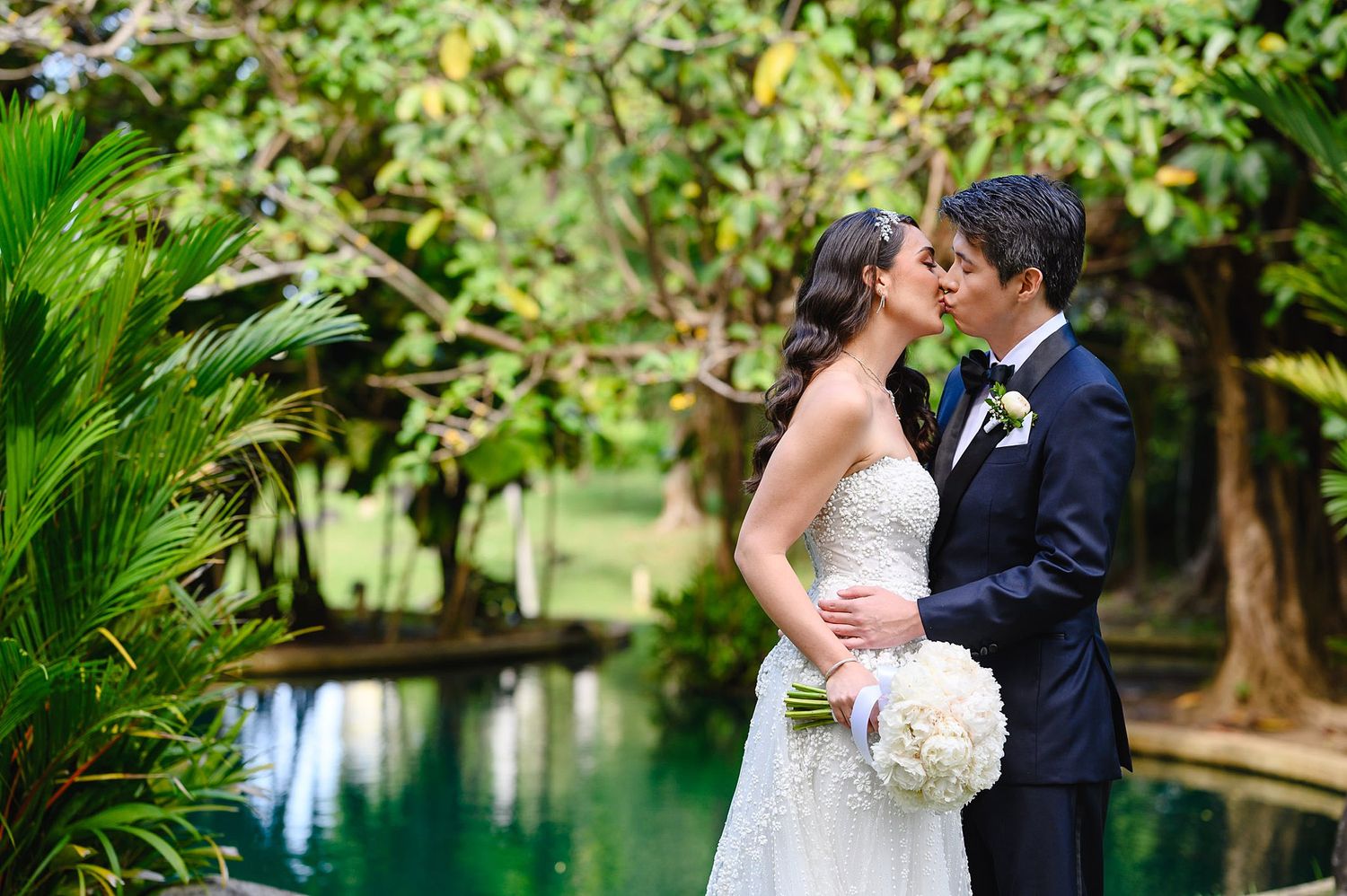 Bride and groom share a romantic kiss in a lush garden setting beside a tranquil pool on their wedding day.