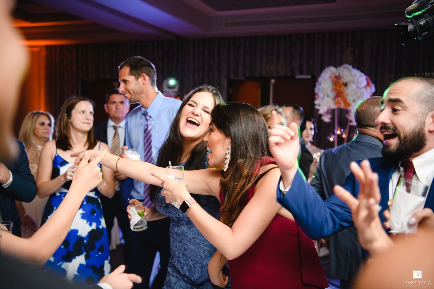 Two women joyfully dancing and embracing at a wedding reception party with guests celebrating in the background.